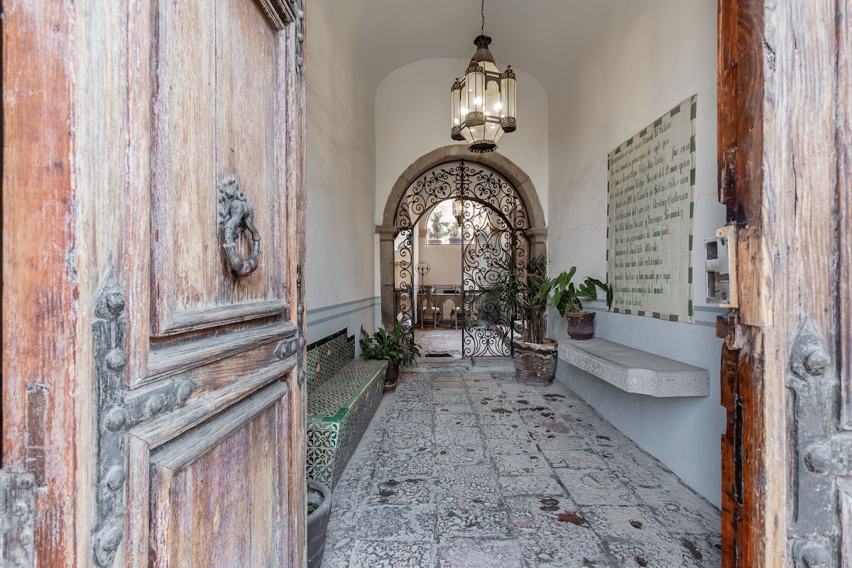 An arched entryway is visible, featuring intricate wrought iron gates set against a backdrop of moonstone-like tiles. The rustic wooden doors have a decorative lion's head knocker, and potted plants are placed along the sides, enhancing the welcoming atmosphere.
