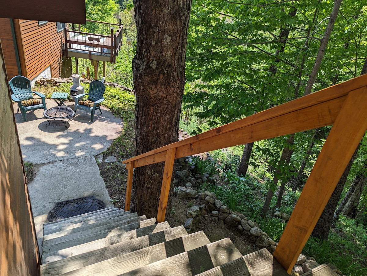 A wooden staircase leads down to a stone patio area surrounded by lush greenery. Two blue chairs and a fire pit are positioned on the patio, offering a cozy outdoor seating space. The setting feels serene, embraced by trees.