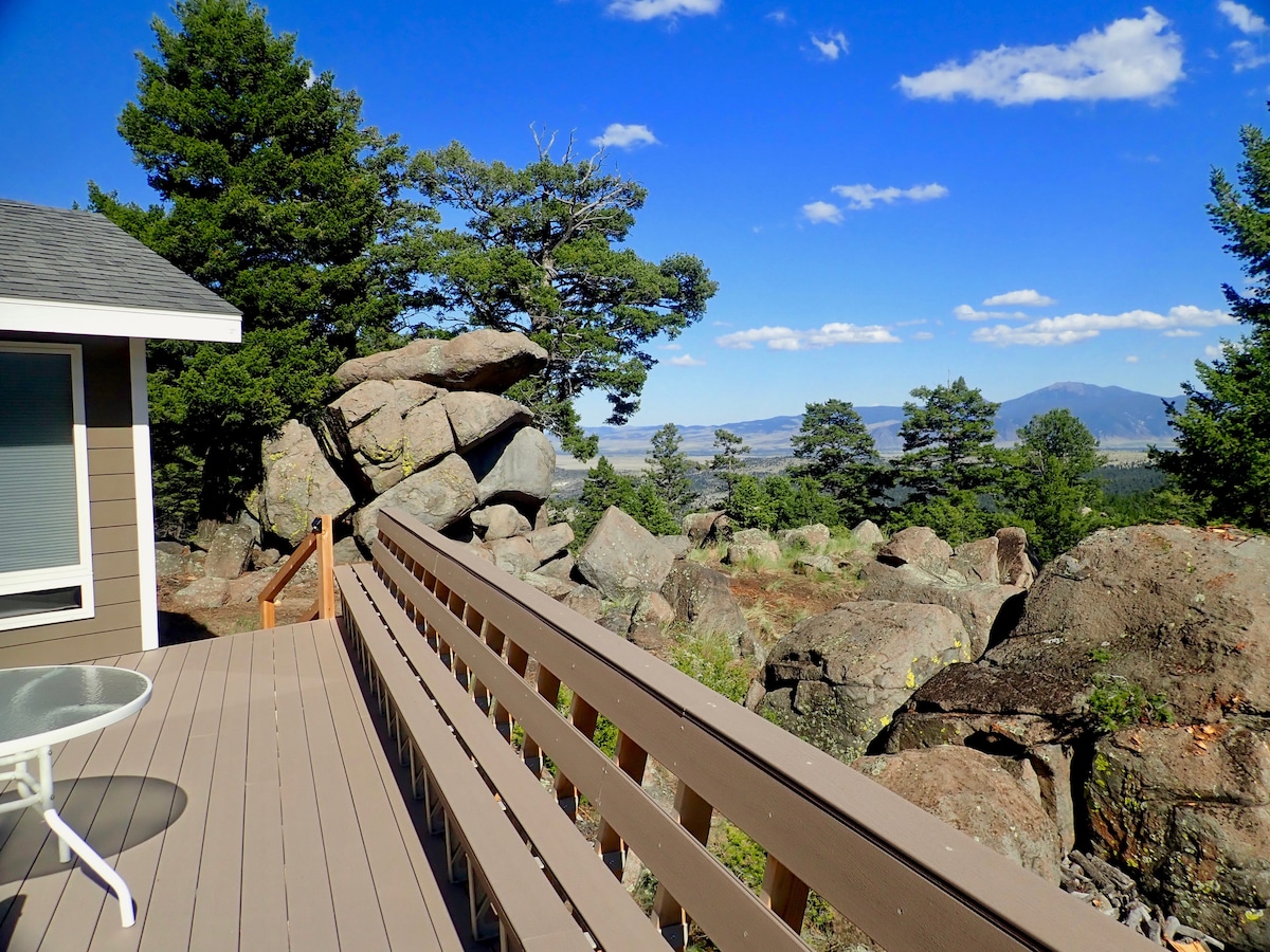 A spacious deck extends from the home, offering serene views of rocky outcrops and verdant trees. A small round table is positioned on the deck, while clear blue skies with scattered clouds provide a peaceful backdrop to the natural landscape.
