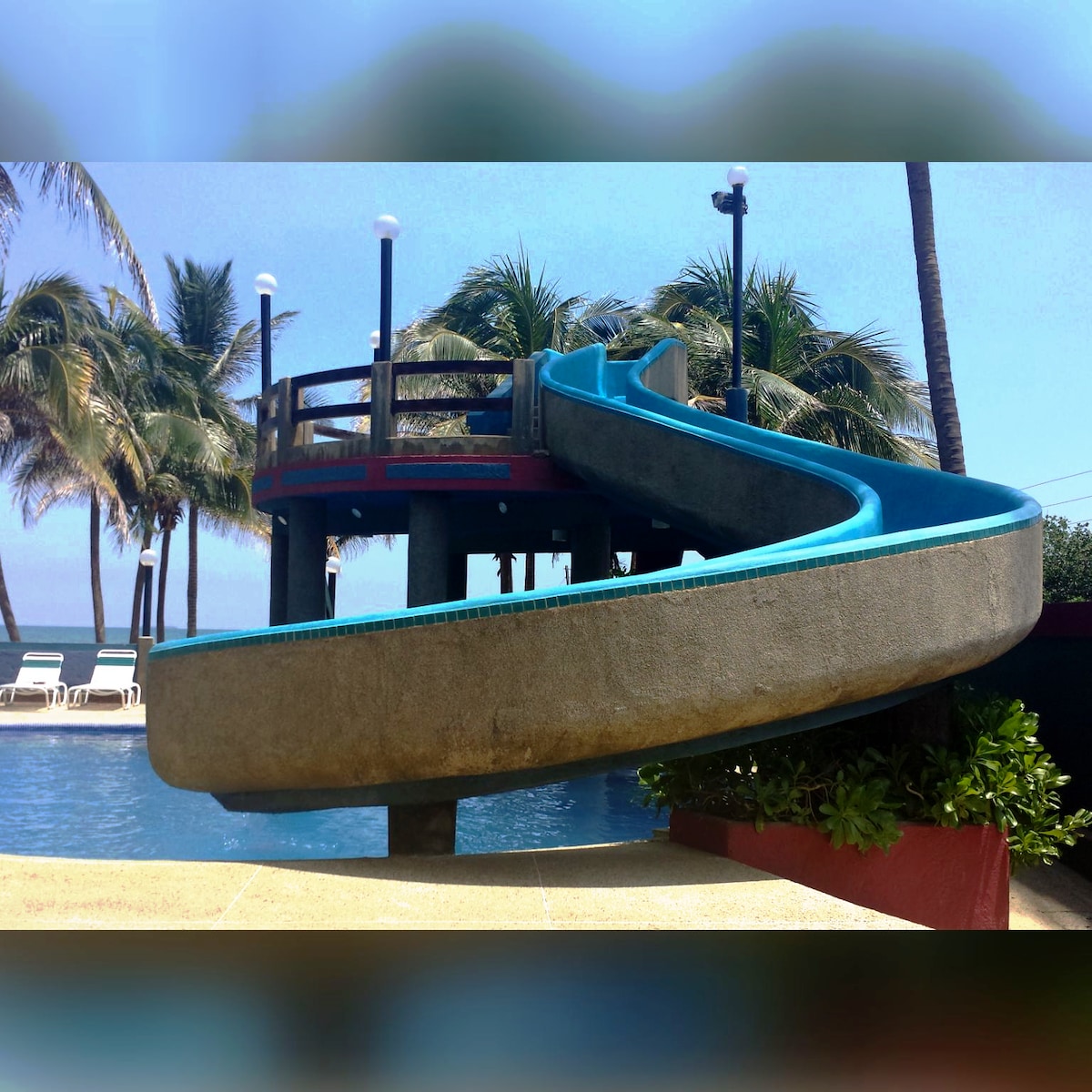 A water slide winds down towards a sparkling pool, surrounded by palm trees. Lounge chairs are positioned nearby for relaxation, and lighting fixtures are installed for evening use. The clear blue sky enhances the vibrant setting, inviting enjoyment and leisure.