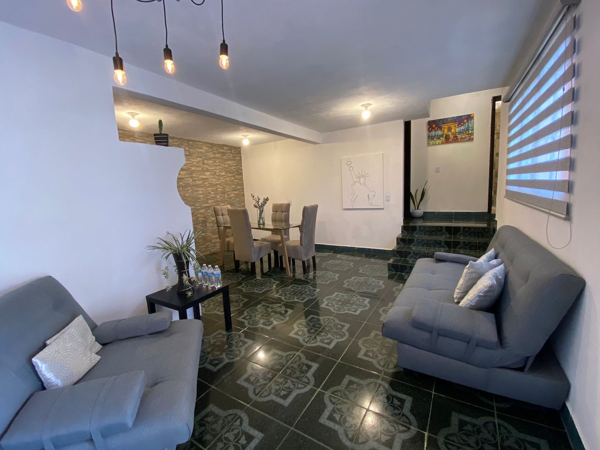 A spacious living area features two gray sofas, a black table with water bottles, and a four-person dining table. Decorative lighting hangs from the ceiling, and the floor is adorned with dark tiles, providing a modern contrast to the light walls and soft furnishings.