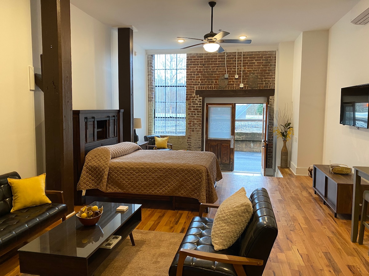 A spacious loft area features exposed brick walls and hardwood flooring. A queen size bed is positioned against one wall, with a simple brown quilt. A cozy seating arrangement includes two black leather chairs and a coffee table, providing a comfortable gathering space.