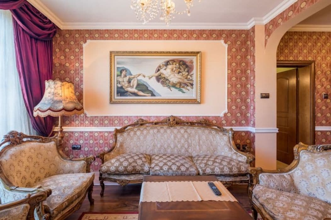 An elegantly furnished living area features ornate seating with plush upholstery, complemented by a decorative chandelier overhead. A classic artwork hangs above a polished wooden table, while rich curtains frame a window, enhancing the baroque ambiance of the space.