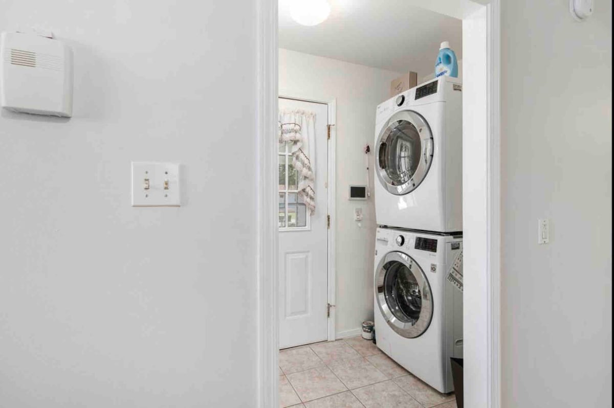 A laundry area features a stackable washer and dryer, equipped with modern controls. The adjacent door leads to the exterior, while neutral-colored walls and tiled flooring create a clean and functional space. Laundry supplies are visible on top of the dryer.