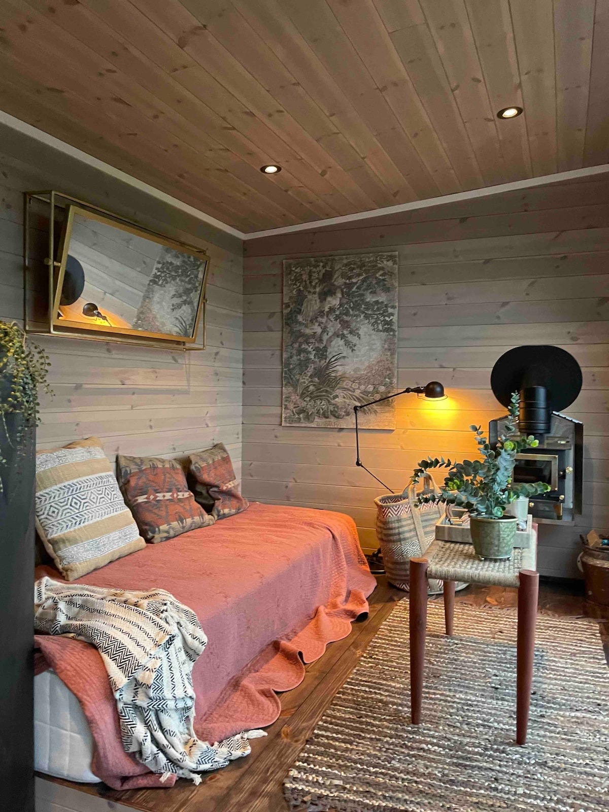 A cozy sitting area features a comfortable sofa adorned with patterned cushions. Natural light filters through the room, illuminating a small table with a potted plant. A stylish lamp and decorative mirror enhance the inviting atmosphere, complemented by wooden paneling and a textured rug.