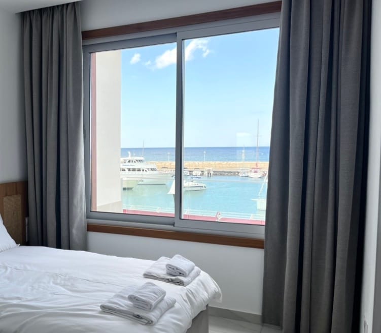 A bright bedroom features a large window offering a view of the marina and sea. Light curtains frame the window, and a neatly made bed is visible with folded towels placed at the foot.
