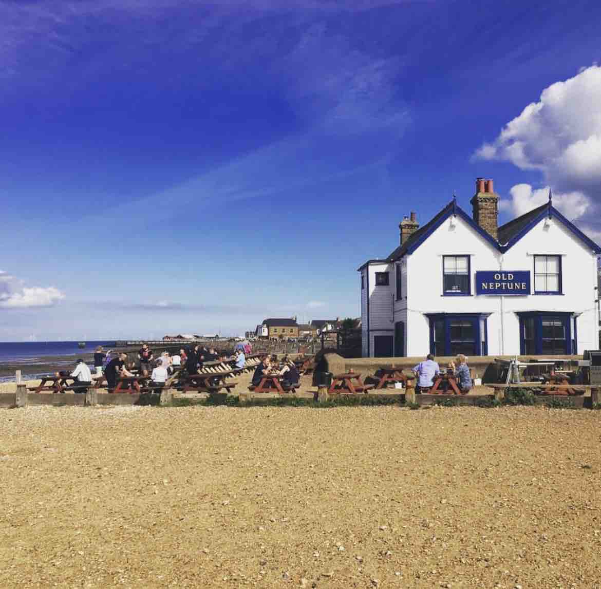 The vibrant beachfront scene captures a traditional white building with blue trim, labeled 'Old Neptune.' Wooden picnic tables are arranged on a sandy area in front, with guests enjoying the outdoor space. The expansive sky is dotted with clouds, and the ocean lies just beyond.