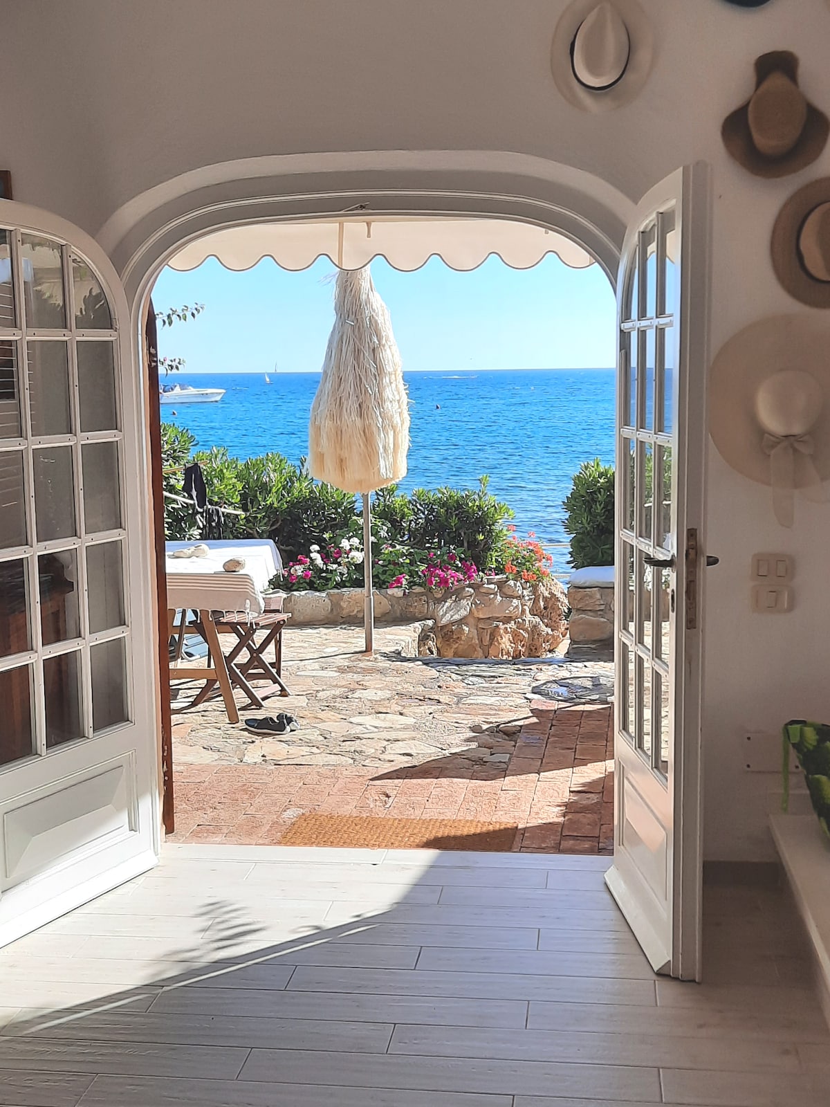 An arched doorway opens to a vibrant outdoor terrace showcasing a stone-paved area adorned with colorful flowers. A simple wooden table and chairs sit under a retractable awning, with the serene sea visible in the background, enhancing the peaceful coastal atmosphere.