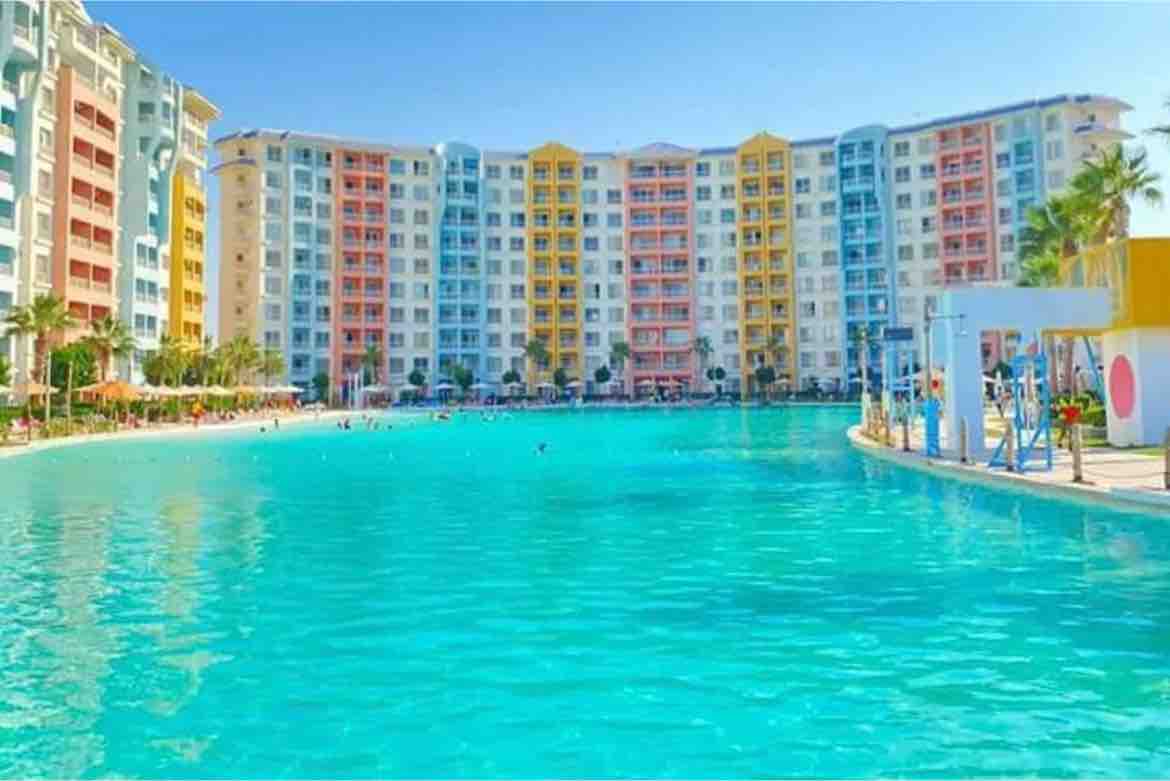 A large swimming pool reflects the bright blue sky, surrounded by colorful, multi-story buildings. Palm trees line the pool's edge, providing shade to sunbathers enjoying the water. Several people can be seen swimming and relaxing in the inviting atmosphere.
