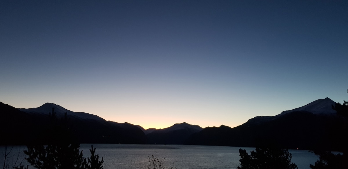 A serene landscape is showcased, featuring a fjord surrounded by mountains. The sky transitions from deep blue to soft hues near the horizon, indicating dusk. The calm water reflects the fading light, enhancing the tranquil ambiance of the natural setting.