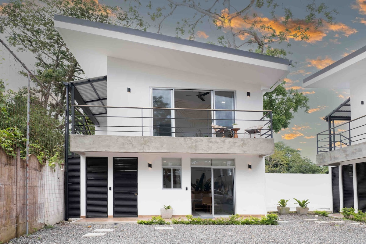 A modern two-story loft building features a sleek exterior with large windows and a balcony. The concrete and white walls are complemented by simple landscaping. A gravel pathway leads to the entrance, offering easy access to the unit.