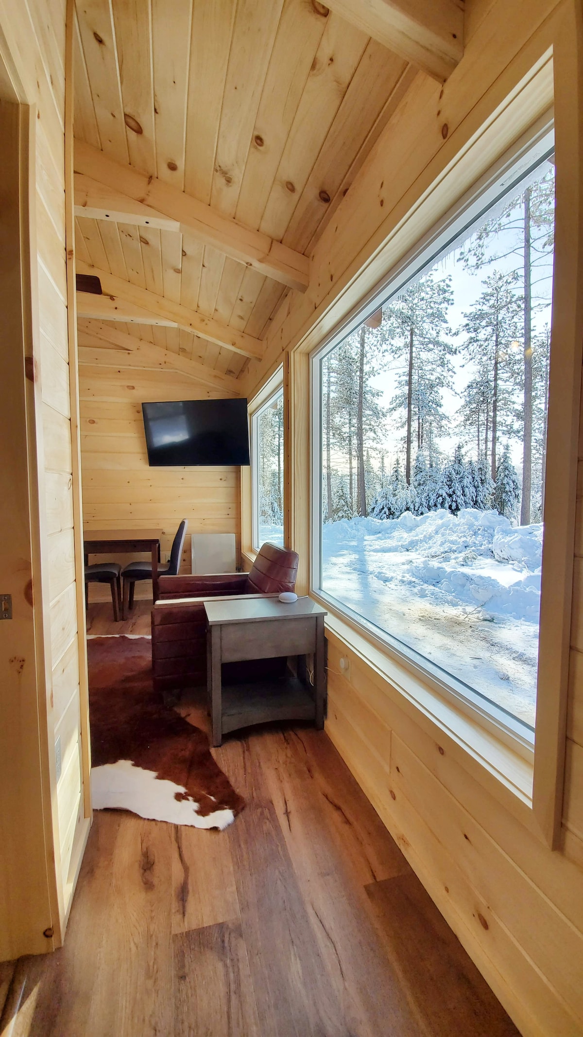 A cozy hallway in the cabin is shown, featuring two large windows that provide views of the snowy landscape and forest. A wooden table and chairs are visible, alongside a small side table. A flat-screen TV is mounted on the wall.