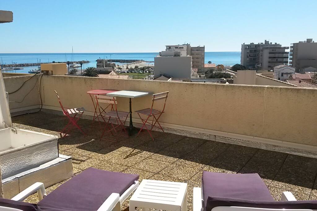 vue mer et port depuis la terrasse toiture avec tables pour manger, chaises (entre 7 et 8) et transats.