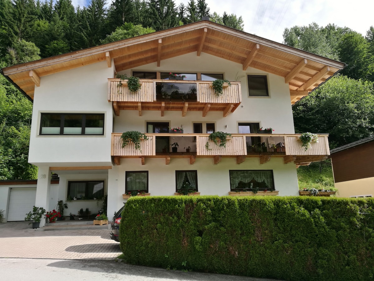 A modern three-story house is set against a backdrop of lush greenery, featuring multiple wooden balconies adorned with potted plants. Large windows allow ample natural light, while the entrance is marked by a neat, landscaped garden with a well-maintained hedge.