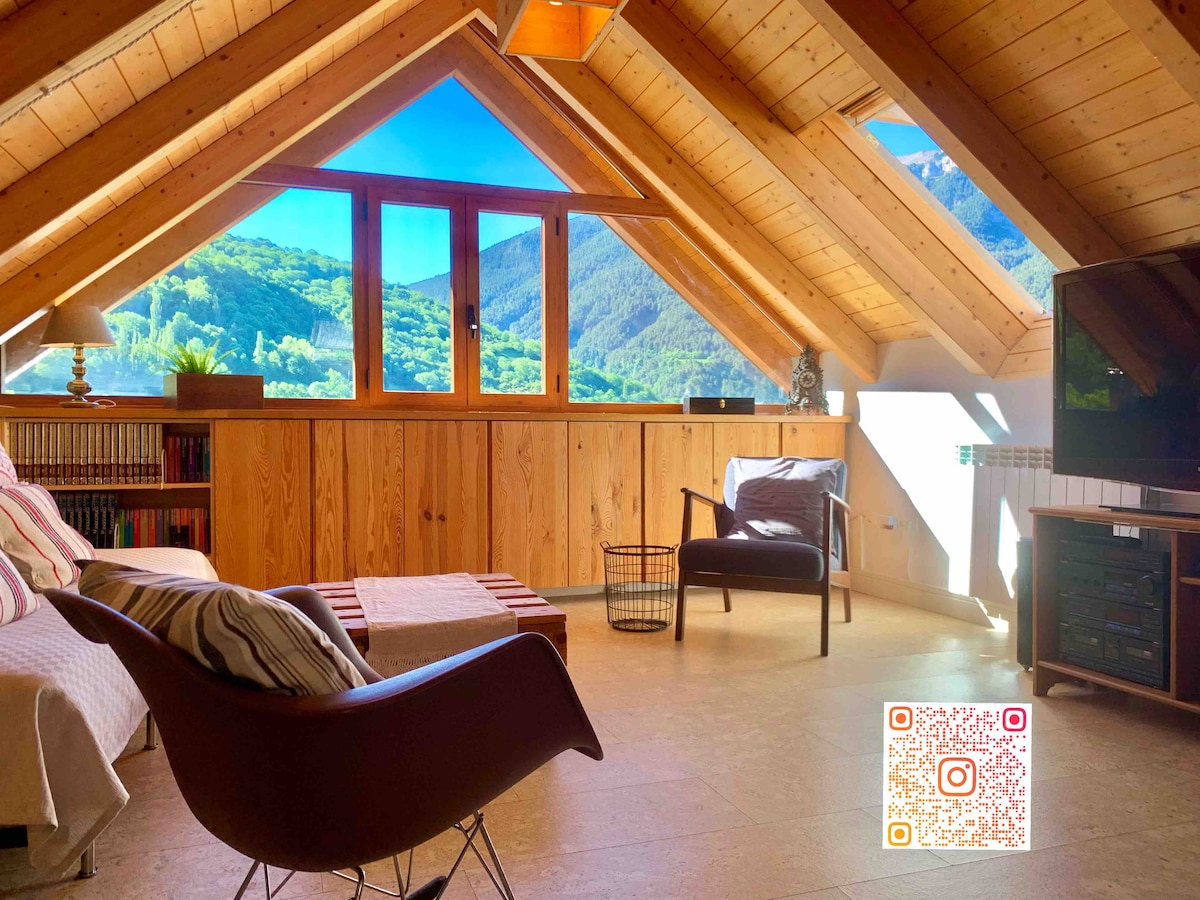 The upper living area features pitched wooden ceilings and large windows that provide sweeping views of the surrounding mountains. A comfortable seating arrangement includes a chair and a sofa, with a wooden coffee table placed centrally. Light floods the space, creating a bright and inviting ambiance.