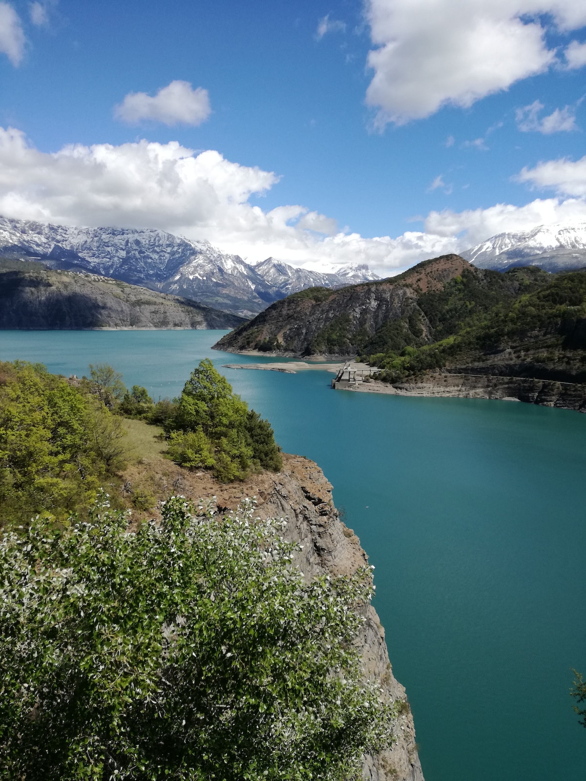 A scenic view of turquoise waters rippling gently against rugged cliffs is presented. Surrounding mountains, capped with snow, rise majestically in the background. Lush greenery frames the foreground, contributing to the peaceful atmosphere of this natural landscape.