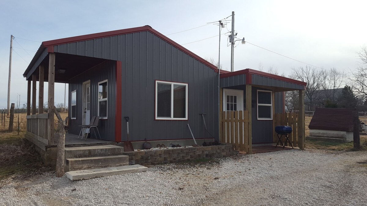 The exterior of a country cabin is displayed, featuring a combination of grey and red siding. A porch with seating is visible, alongside a gravel driveway. A small fenced area, visible on the right, adds a rustic touch, while utility poles are included in the background.
