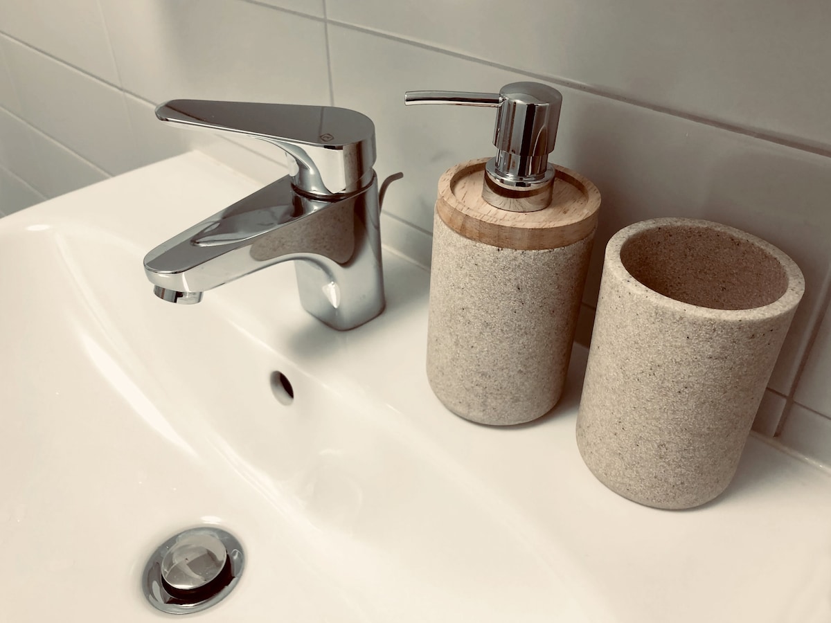 A modern sink area is displayed, featuring a sleek chrome faucet alongside a soap dispenser with a wooden accent and a matching stone cup for toiletries. The white countertop complements the clean lines of the ceramic sink, enhancing the space's contemporary feel.