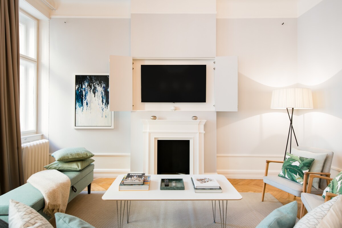 A stylish living room features a wall-mounted TV above a white fireplace. A modern coffee table sits on a beige area rug, surrounded by a comfortable sofa and a blue accent chair. Natural light enters through large windows with soft curtains, complemented by abstract art on the wall.