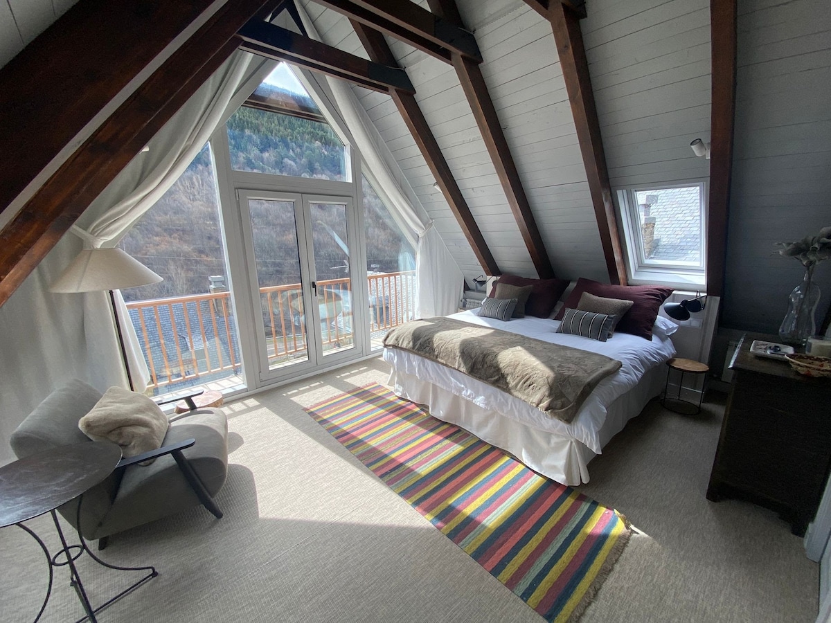 A bright and spacious bedroom features a sloped ceiling with wooden beams. A large bed is dressed in soft textiles, and natural light streams through glass doors leading to a private balcony. A colorful area rug adds warmth, complemented by a small seating area.