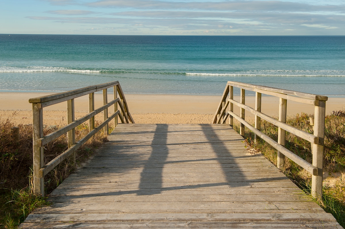 A wooden pathway leads to an expansive sandy beach, with gentle waves lapping against the shore. The clear blue water extends to the horizon, and soft clouds are visible in the sky, providing a serene coastal atmosphere.