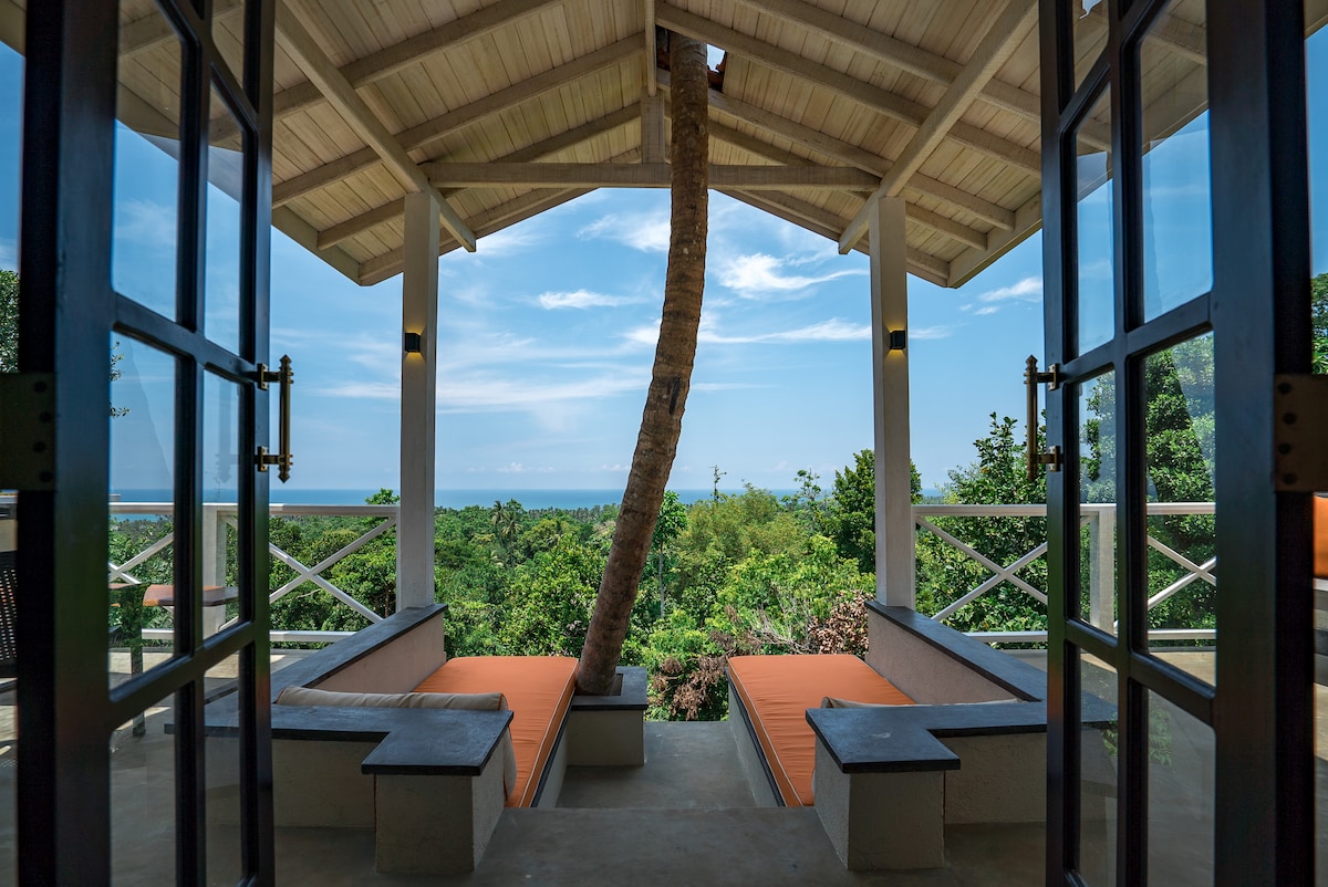 Large glass doors open to a spacious outdoor seating area, featuring wooden benches with comfortable cushions. A palm tree is visible in the foreground, framed by lush greenery and a panoramic view of the coastline and ocean beyond.
