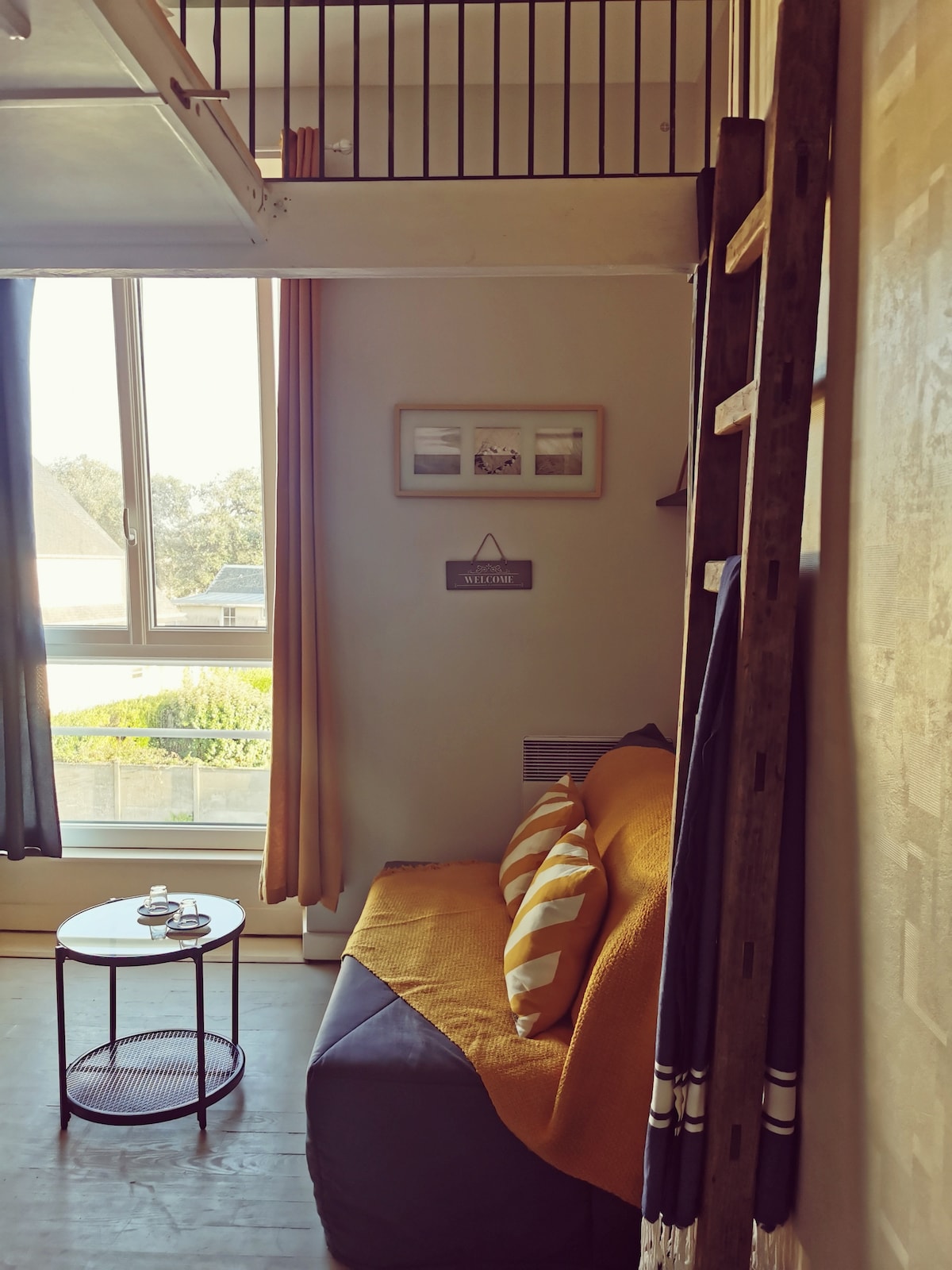 A cozy seating area is featured with a dark-colored sofa adorned with striped cushions. A small round table sits nearby, holding a couple of items. Natural light filters through a large window, complemented by flowing curtains, providing a serene view of the garden.