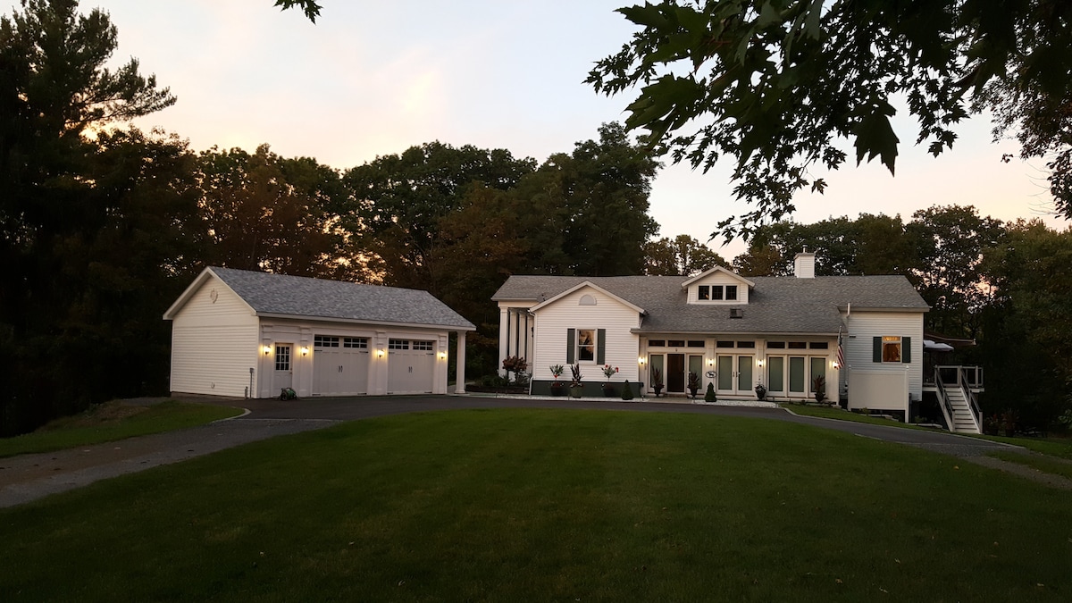 A spacious home is shown, featuring elegant architecture and a well-maintained lawn. A driveway leads to an attached garage, while planters with greenery add a touch of color along the entrance. A serene natural backdrop of trees enhances the setting.