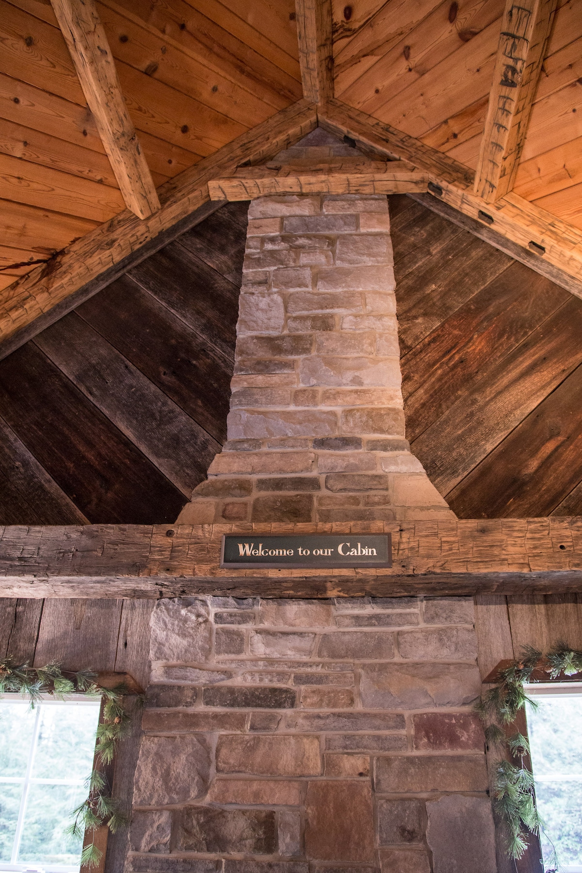 The interior features a tall stone chimney, constructed from various sized stones, rising to a wooden peak. Wooden beams accentuate the ceiling, and a sign reading 'Welcome to our Cabin' adds a personal touch. Windows allow natural light to illuminate the space.