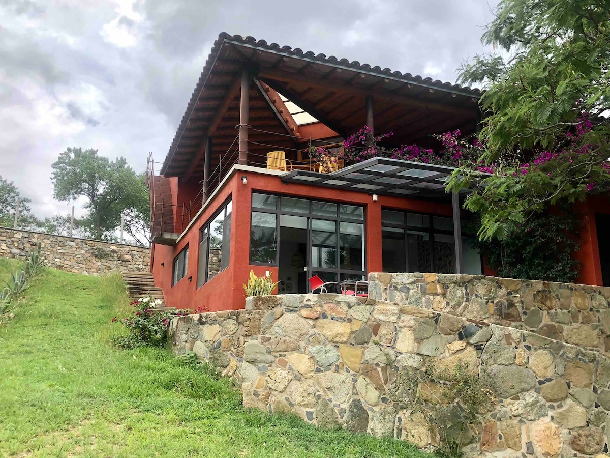 A modern building with a red exterior features large glass windows and a spacious terrace adorned with flowering plants. The stone foundation and surrounding greenery complement the natural landscape, creating a harmonious connection to the outdoors.