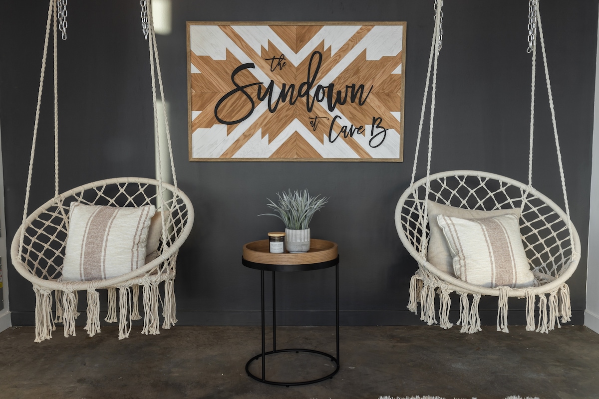 Two hanging chairs are positioned side by side, showcasing a modern design with fringe details. A round wooden table sits between them, adorned with a small potted plant and a decorative cup. A striking wall art piece featuring the name 'The Sundown at Cave B' adds character to the space.