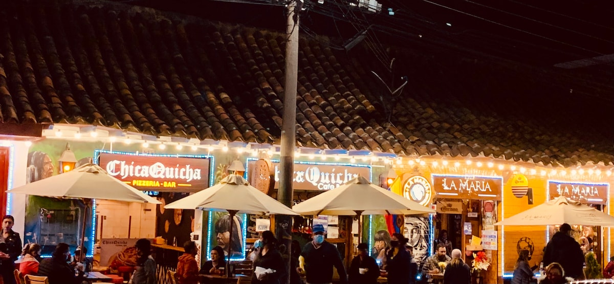 A lively street scene at night is illuminated by colorful signage and string lights. Outdoor dining areas are equipped with large umbrellas, while people are gathered around tables, enjoying the ambiance. The rustic tiled roof above adds character to the vibrant atmosphere.