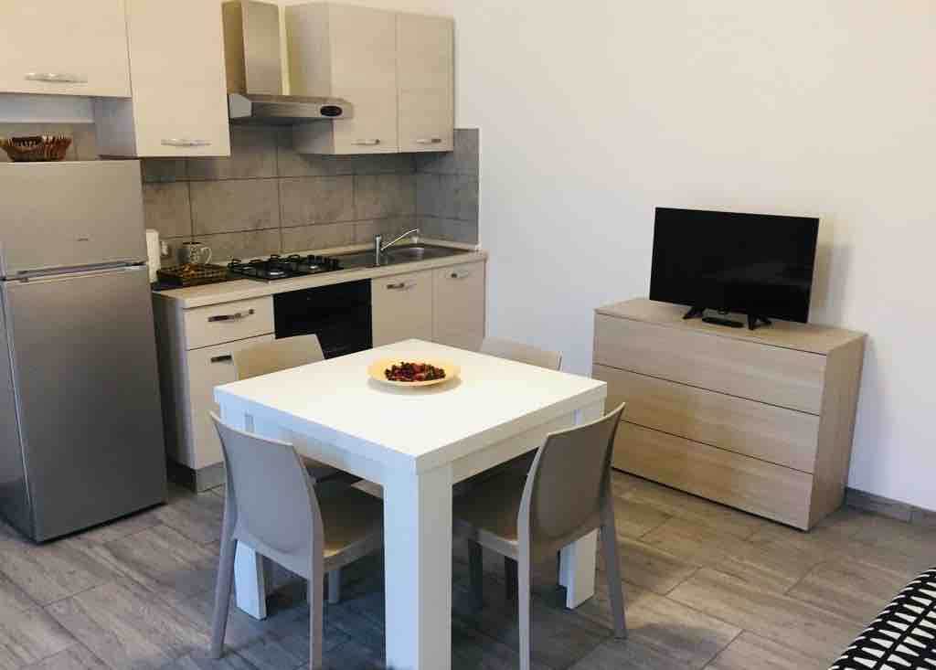 A compact kitchen area is visible, featuring modern cabinetry and appliances, including a refrigerator and stove. A dining table with four chairs is positioned nearby. Natural light fills the space, highlighting the neutral tones of the furnishings and the tiled floor.