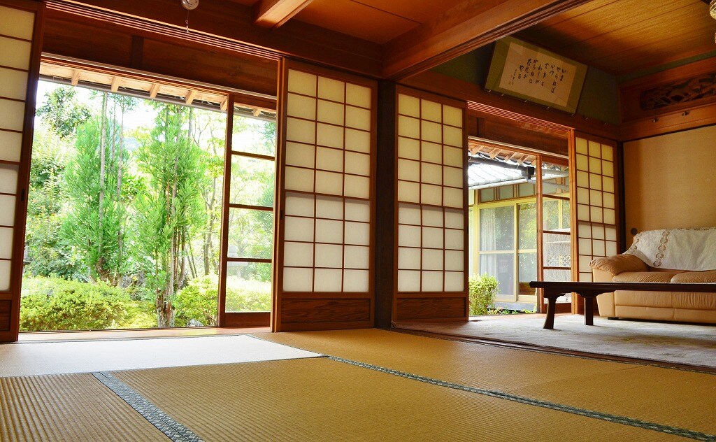 A traditional tatami room features shoji doors that open to a serene garden. Natural light filters through the sliding doors, illuminating the wooden decor. A low coffee table is positioned on the tatami mat, offering a simple and peaceful setting.