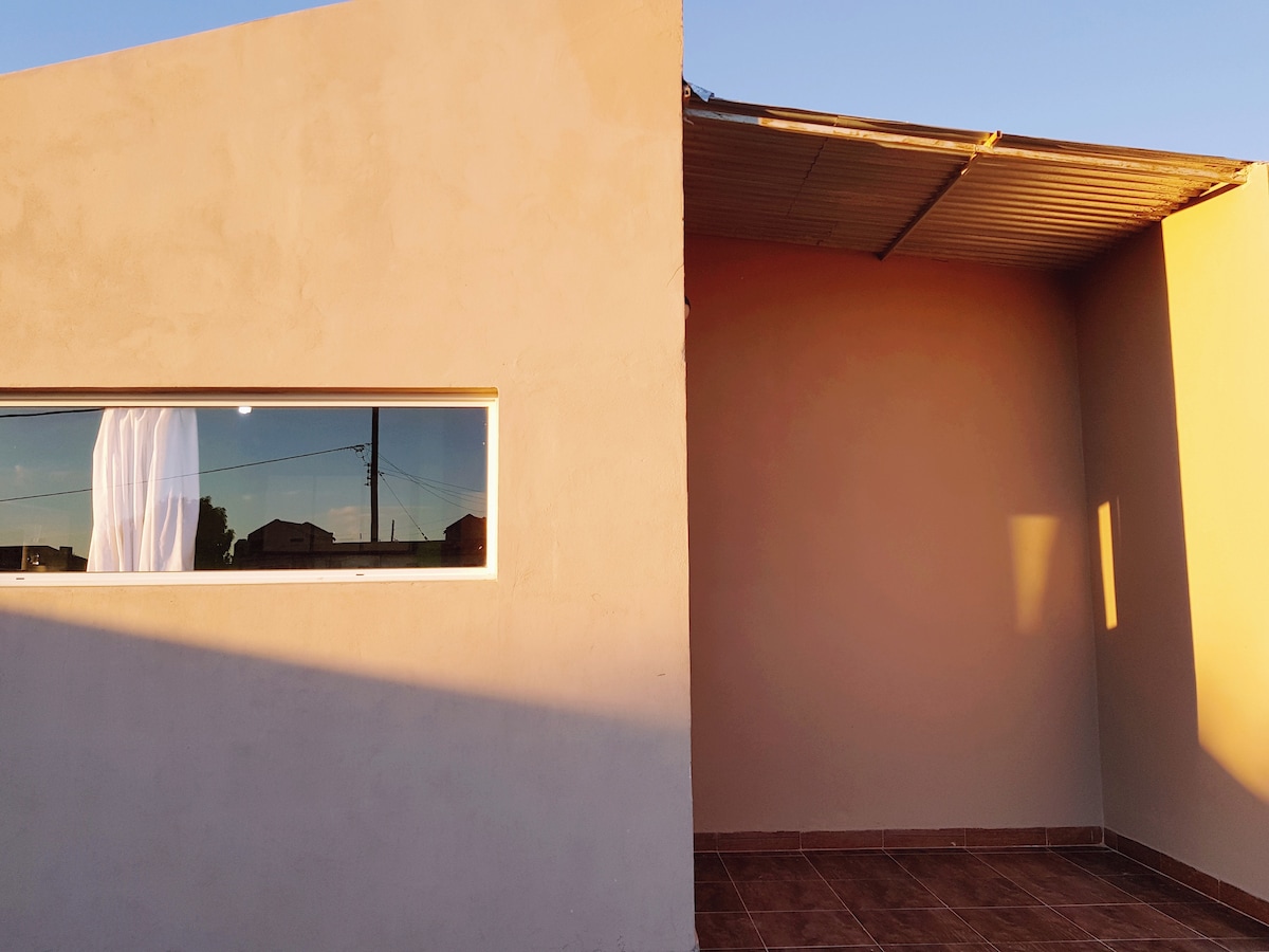 An exterior view of the building is depicted, showcasing a simple architectural design. A large window allows natural light to enter, with sheer white curtains softly swaying. The area is surrounded by a clean, neutral-colored wall and a tiled patio with ample space for relaxation.