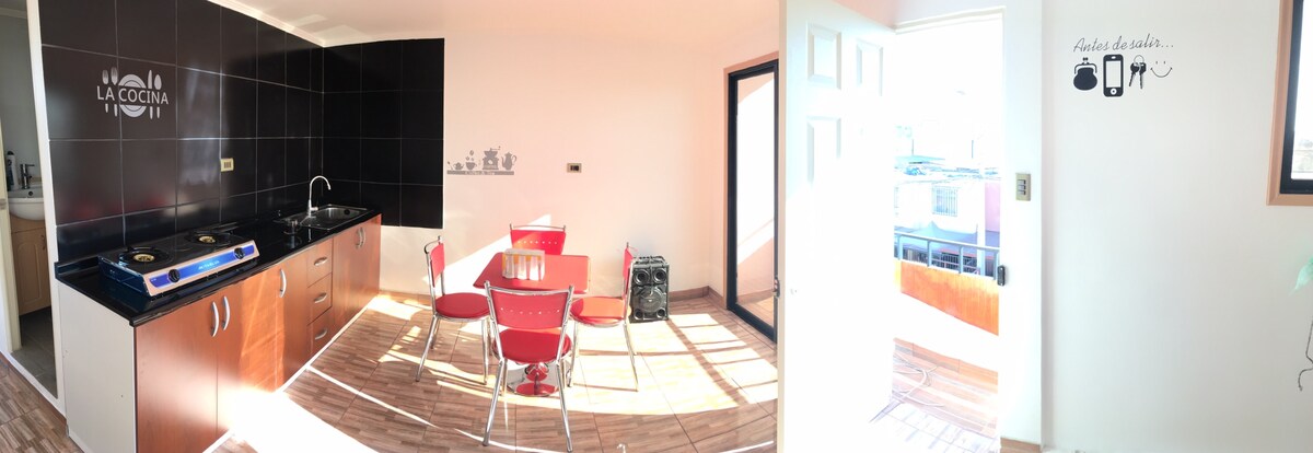 A spacious kitchen and dining area is presented with a modern design. A round table with four red chairs is centrally located, while a black backsplash contrasts with the light cabinetry. A doorway leads to the outside, allowing natural light to fill the space.