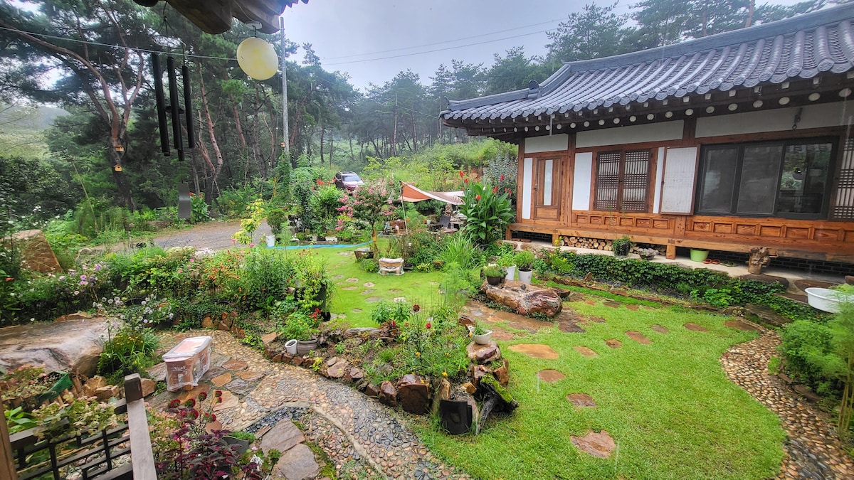 A serene garden surrounds a traditional Korean house, featuring vibrant flowers and neatly arranged stones. Lush greenery and a gentle pathway enhance the tranquil atmosphere. Nearby, a small vehicle is parked under shaded trees, adding to the peaceful rural setting.