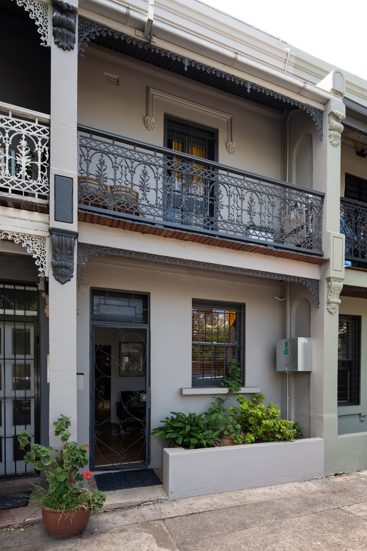 The exterior of a renovated terrace house showcases decorative ironwork balconies and a welcoming entrance. Potted plants line the entryway, enhancing the lush appearance. Soft natural light highlights the building's features, reflecting a blend of classic and contemporary design.