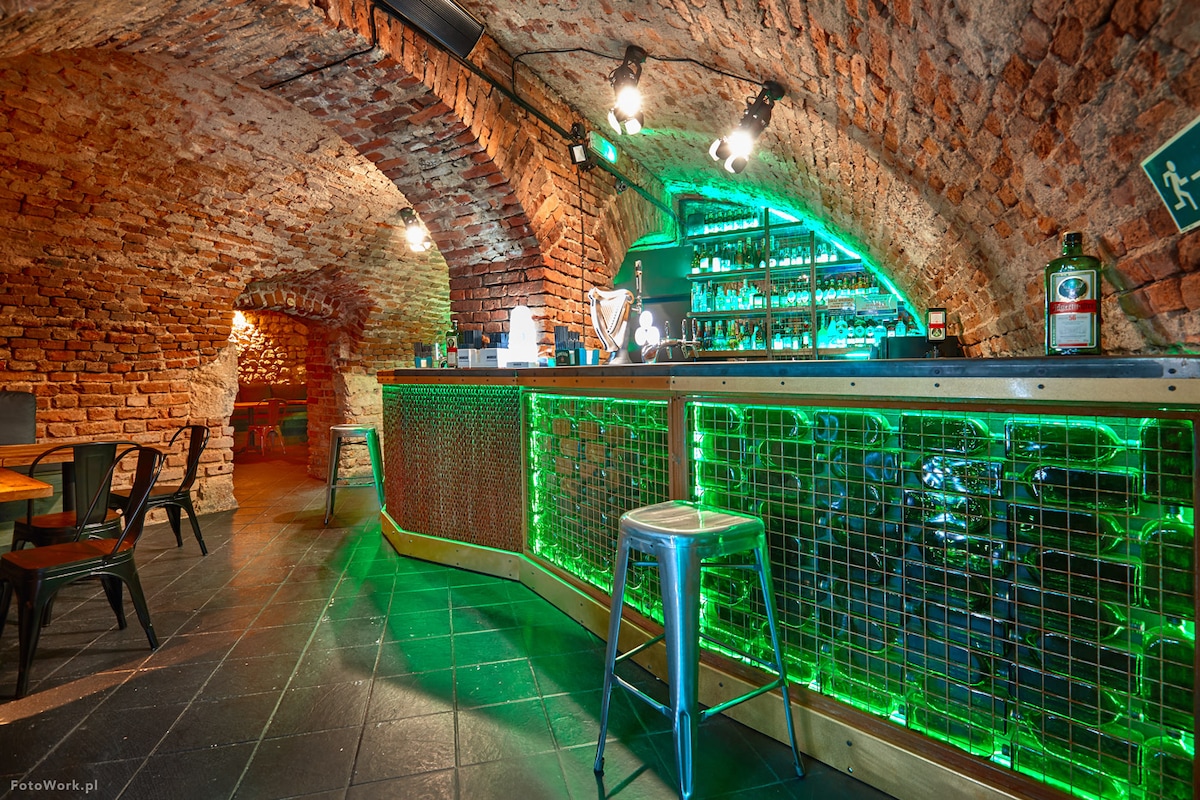 A stylish bar area is showcased within a brick archway, featuring green LED lighting that highlights the counter and shelves stocked with various bottles. Metal stools are positioned at the bar, and the textured walls add to the unique ambiance of the space.
