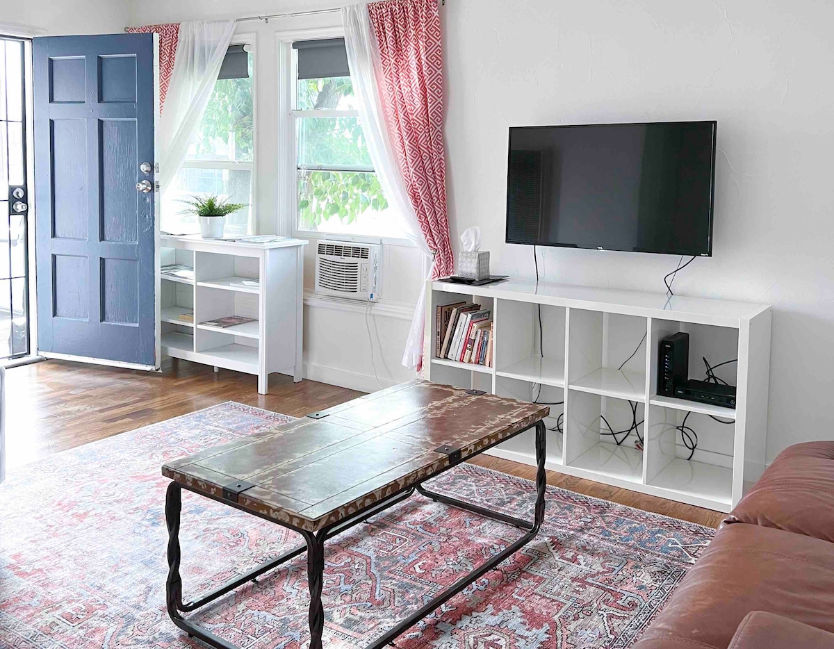 The living area features a comfortable reclining love seat and a coffee table set on a decorative area rug. A mounted television is positioned on a white unit, while large windows with light curtains allow natural light to brighten the space. A separate entrance is visible.