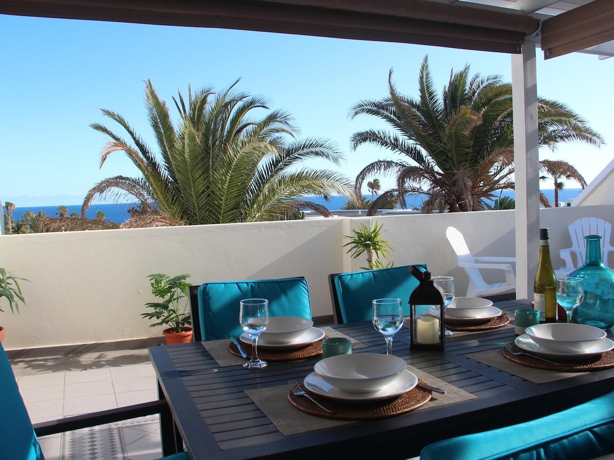 An outdoor dining area is presented with a table set for meals, featuring turquoise chairs and plates. Lush palm trees and ocean views create a serene backdrop, while sunlight illuminates the space, enhancing the inviting atmosphere.