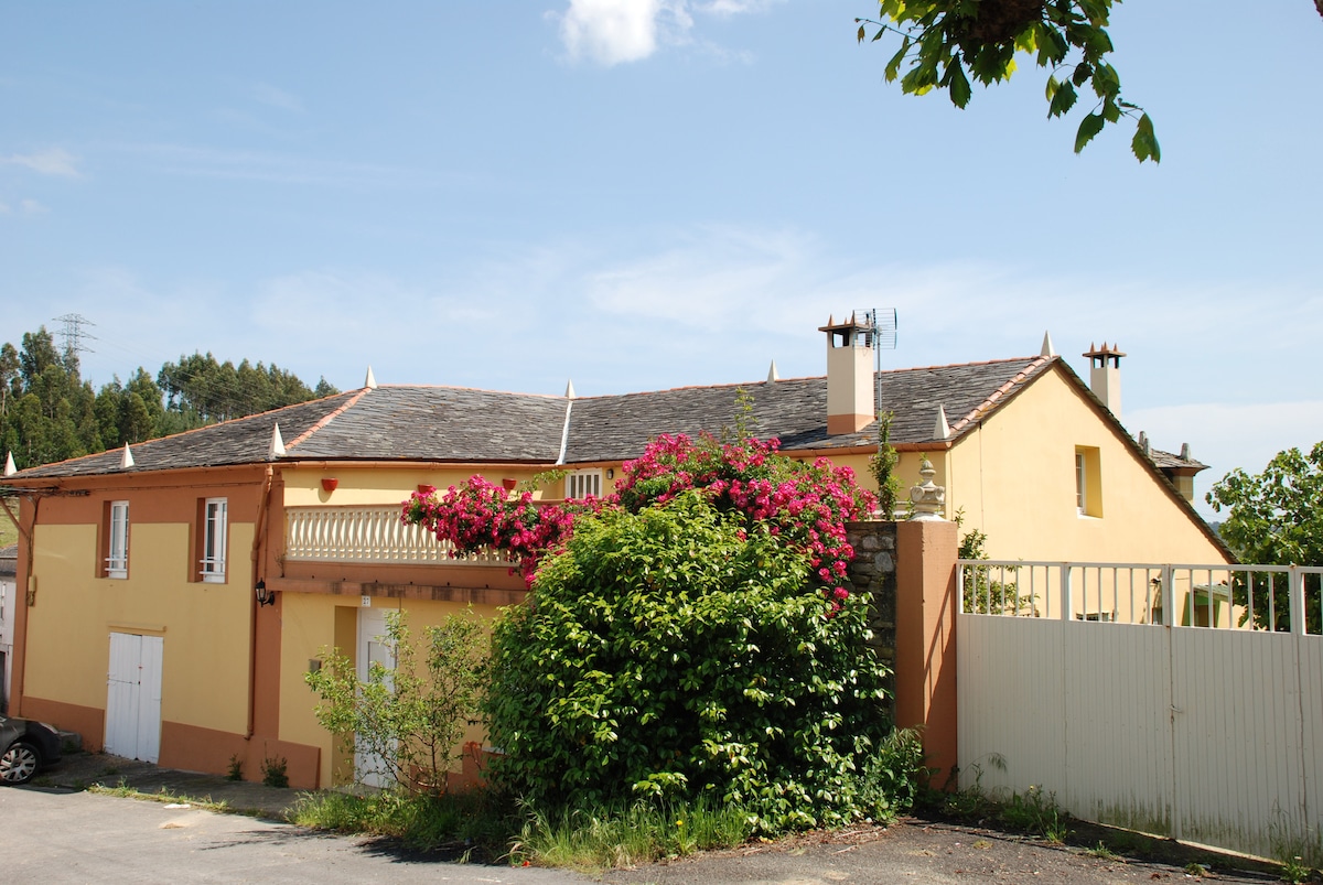 A rural apartment building is surrounded by lush green foliage and blooming flowers. The exterior features soft yellow walls and a sloped roof, with a balcony adorned with climbing plants. A communal parking area is available in front, minimizing disturbances from noise.