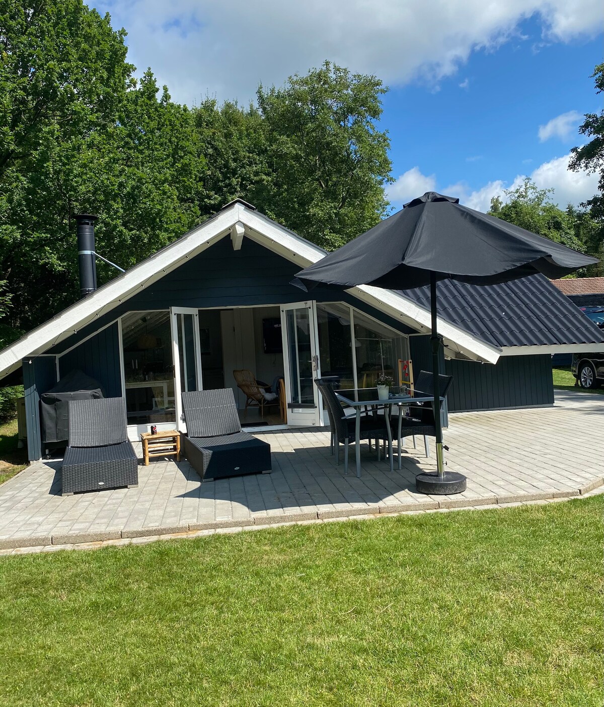 The summer house features a sloped roof and large glass doors opening to a sunlit terrace. A black umbrella shades a dining table and chairs, while two lounge chairs invite relaxation. Lush greenery surrounds the outdoor space, creating a serene environment.