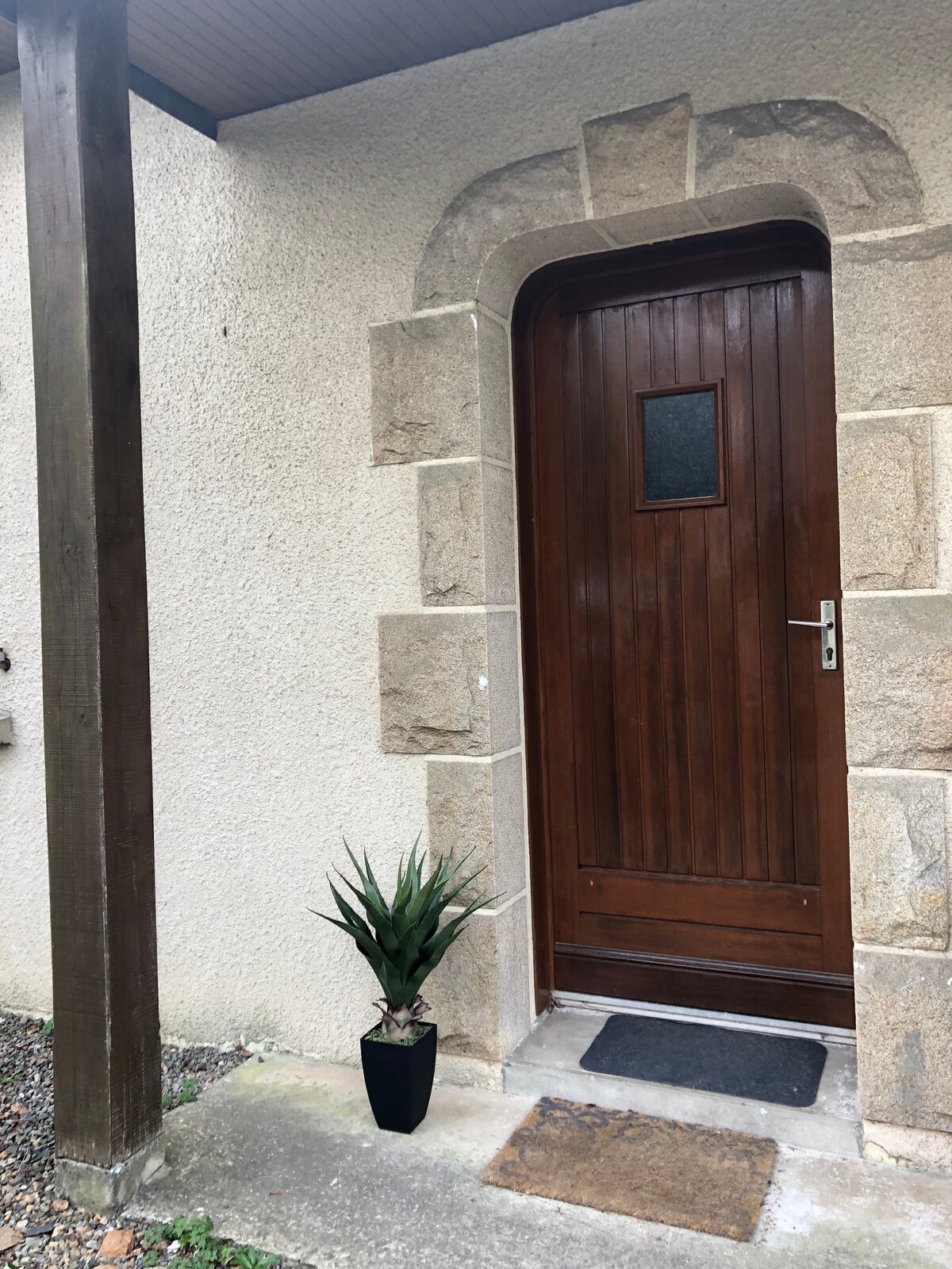 A wooden door with a central window is framed by a stone archway, presenting a welcoming entrance. A small potted plant is positioned beside the door on the concrete step, complementing the earthy tones of the exterior wall.