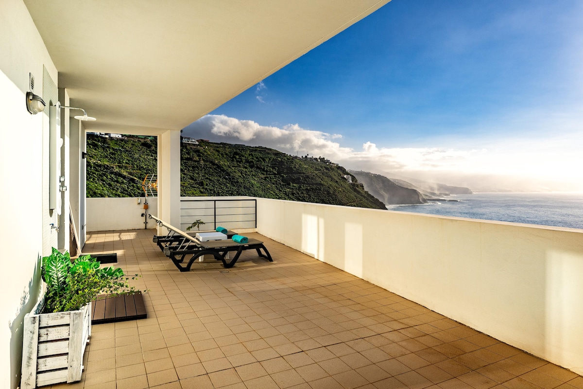 A spacious terrace extends from the villa, featuring sun loungers positioned for optimum views of the coastline and ocean. The backdrop includes lush green hills and a clear sky, providing a serene outdoor setting.