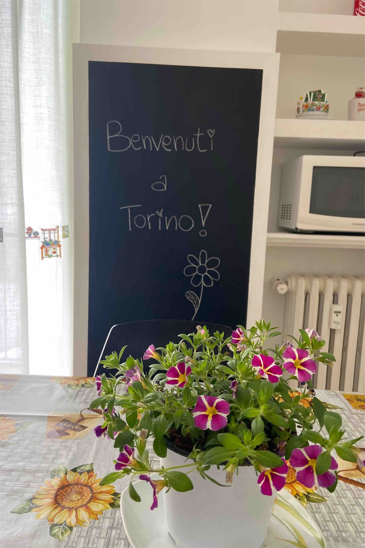 A welcoming kitchen space is adorned with a small potted plant featuring pink and white flowers. A chalkboard displays the message, 'Benvenuti a Torino!' A table is set with a floral tablecloth, while a microwave and a radiator are visible in the background.
