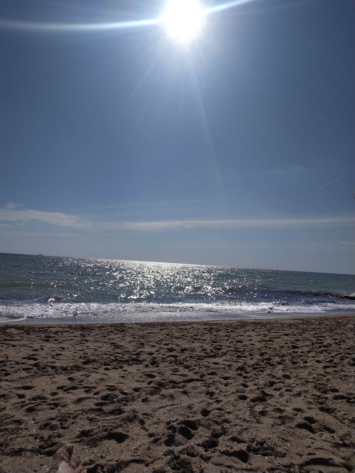 A sandy beach is depicted, with gentle waves lapping at the shore. The sun is situated high in the blue sky, reflecting off the water's surface, creating a shimmering effect. The shoreline is clear and spacious, inviting enjoyment of the coastal setting.