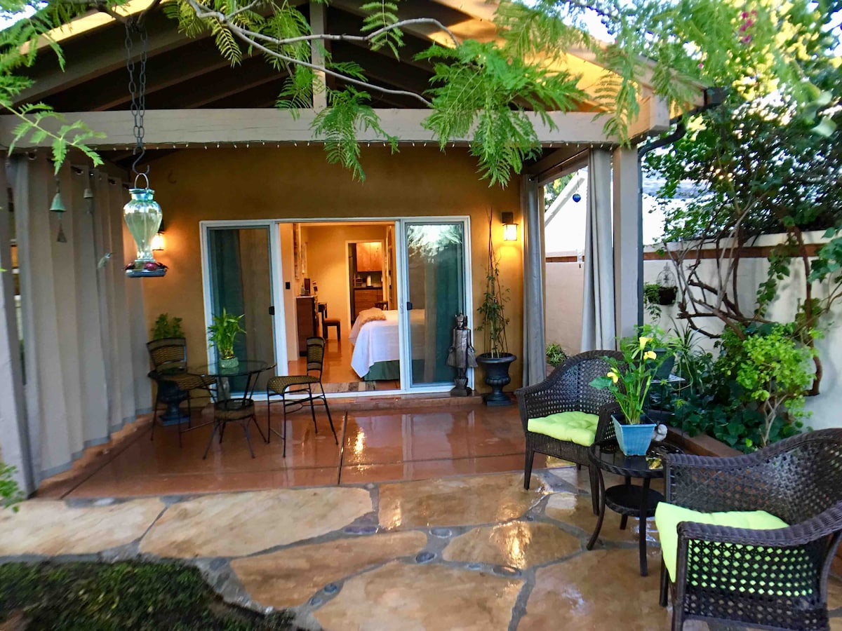 A private patio is framed by lush greenery and features a stone floor. Two wicker chairs with bright cushions and a small table invite relaxation. Sliding glass doors lead into the studio, allowing natural light to illuminate the space.