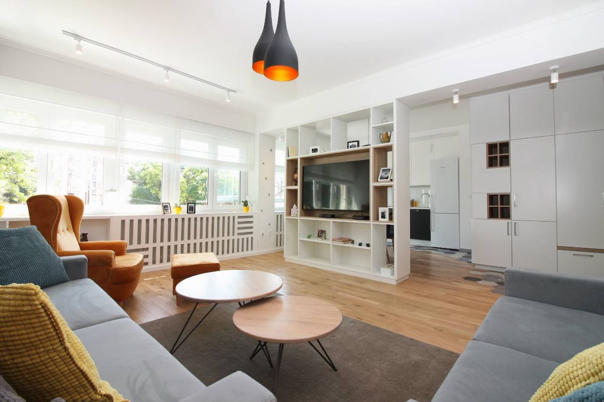 A modern living area features a large window that allows natural light to fill the space. Two orange armchairs are positioned alongside a low coffee table, surrounded by a gray sectional sofa. A mounted flat-screen TV is integrated into a minimalist shelving unit against one wall.