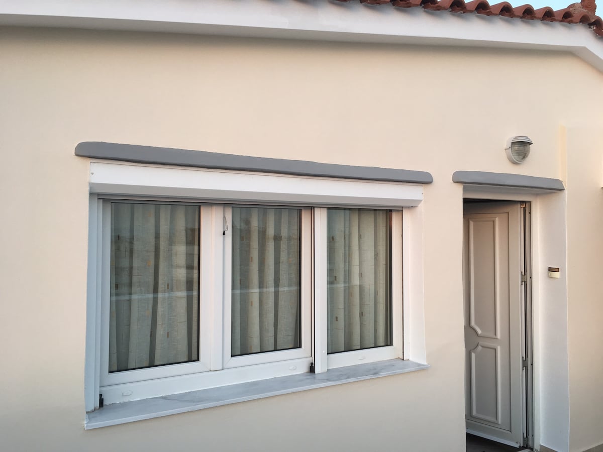 The exterior of the studio features a well-maintained light-colored facade with a tiled roof. A clear entrance door leads into the studio, flanked by large windows with sheer curtains, allowing natural light to filter inside.